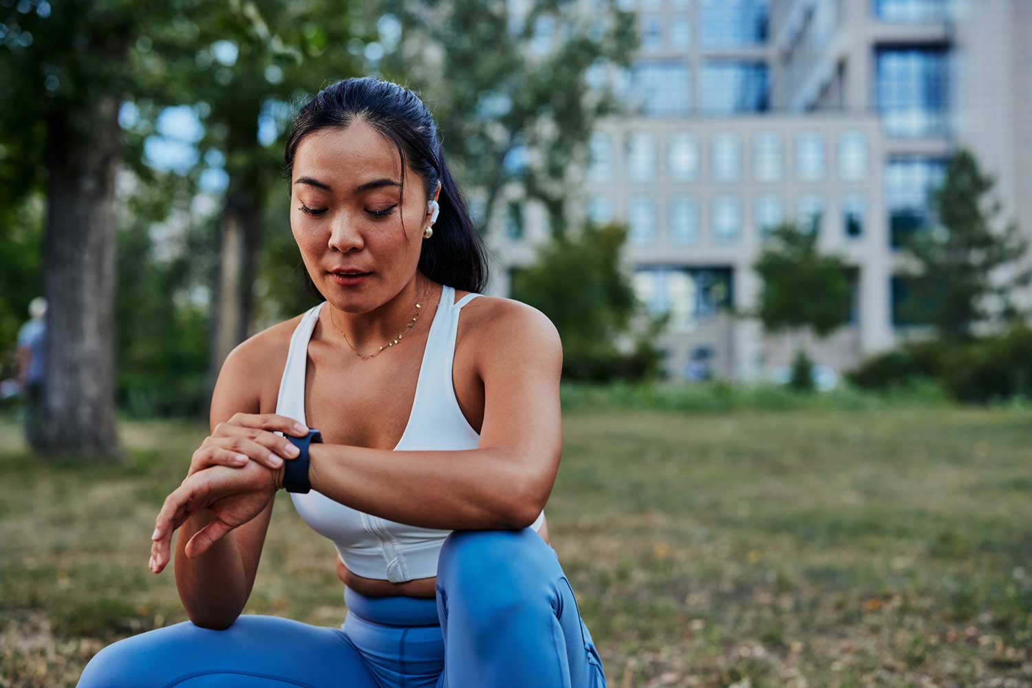 A photograph of a person wearing workout clothes in a park kneeling down to check their smart watch.